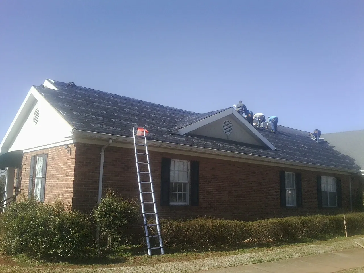 Crew tearing shingles off a brick ranch roof with a ladder set — Metal Roof Repair work by our crew in Holly Springs